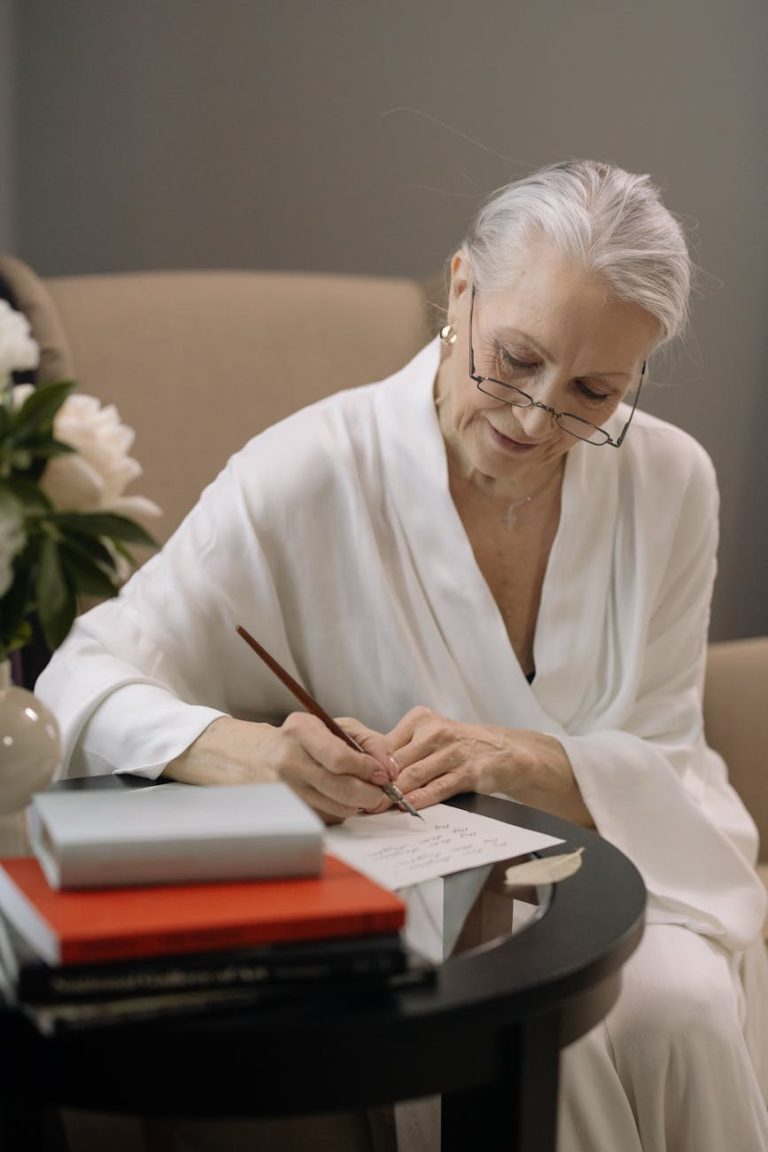Senior woman in eyeglasses writing notes indoors, wearing a white robe. Elegant and focused mood.