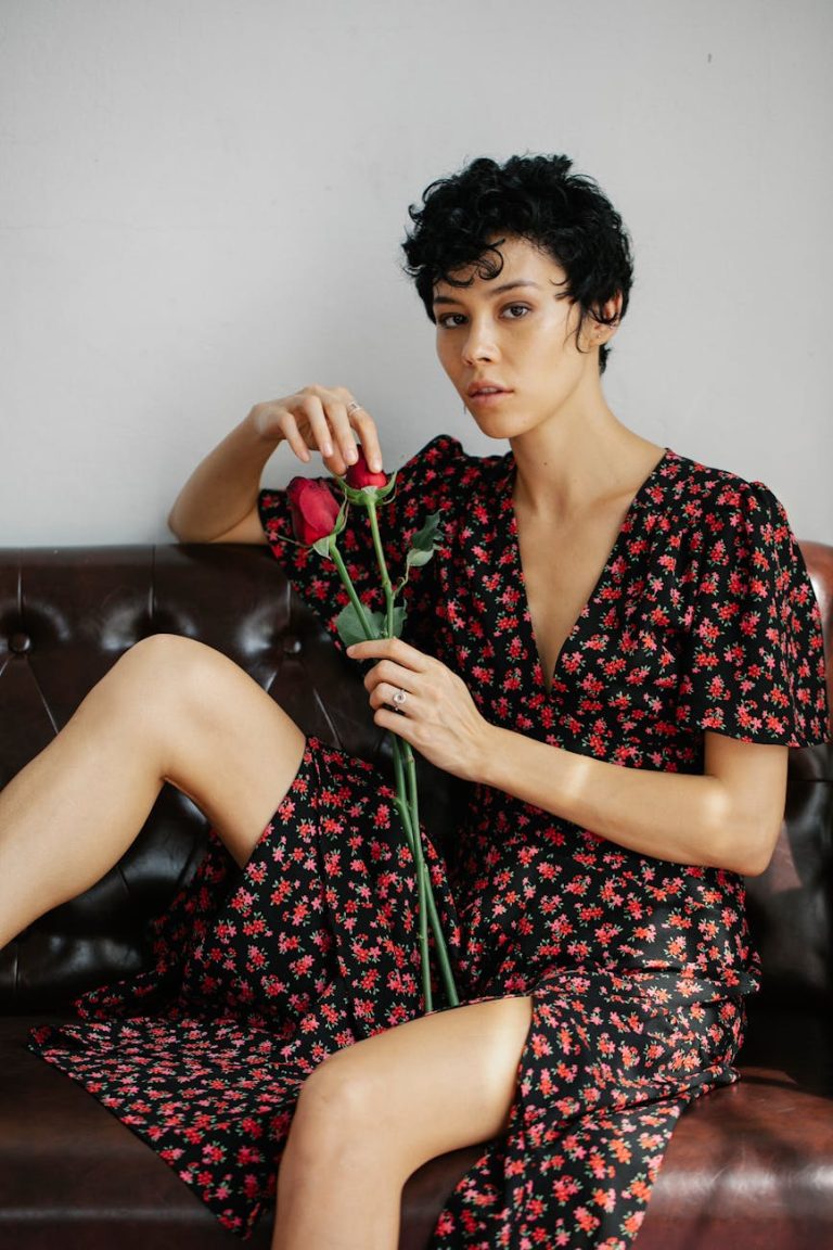 Portrait of a woman in a floral dress holding a red rose on a leather sofa, indoors.