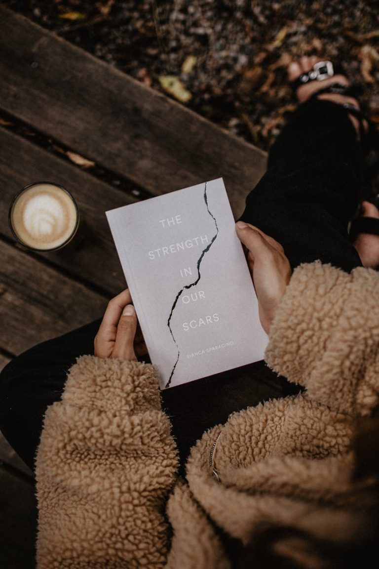 Woman reading 'The Strength in Our Scars' book outdoors in autumn with coffee.