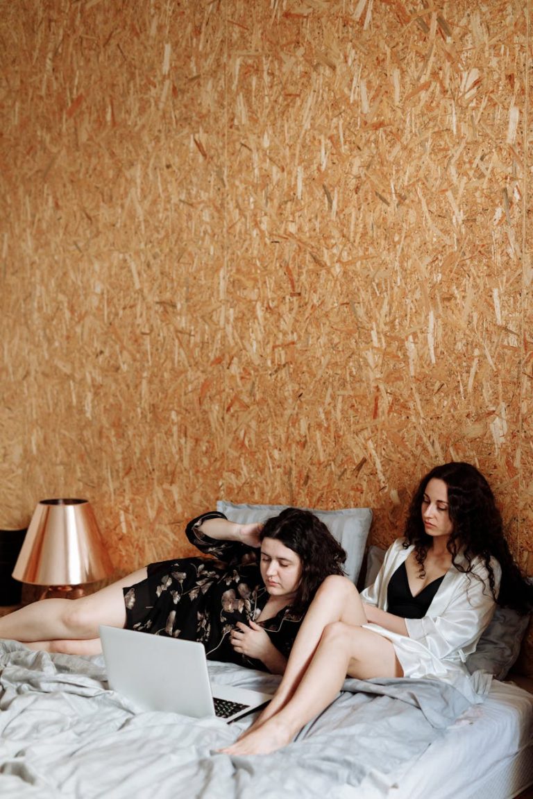 Two women lounging on a bed with a laptop, embracing a calm and intimate atmosphere indoors.