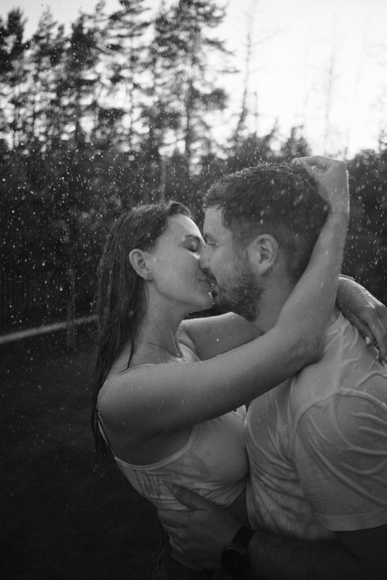 A couple shares a passionate kiss outdoors in the rain, set against a forest backdrop.