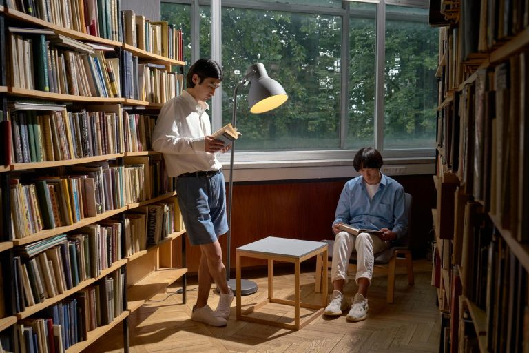 Two men reading in a cozy library setting, surrounded by bookshelves and natural light.