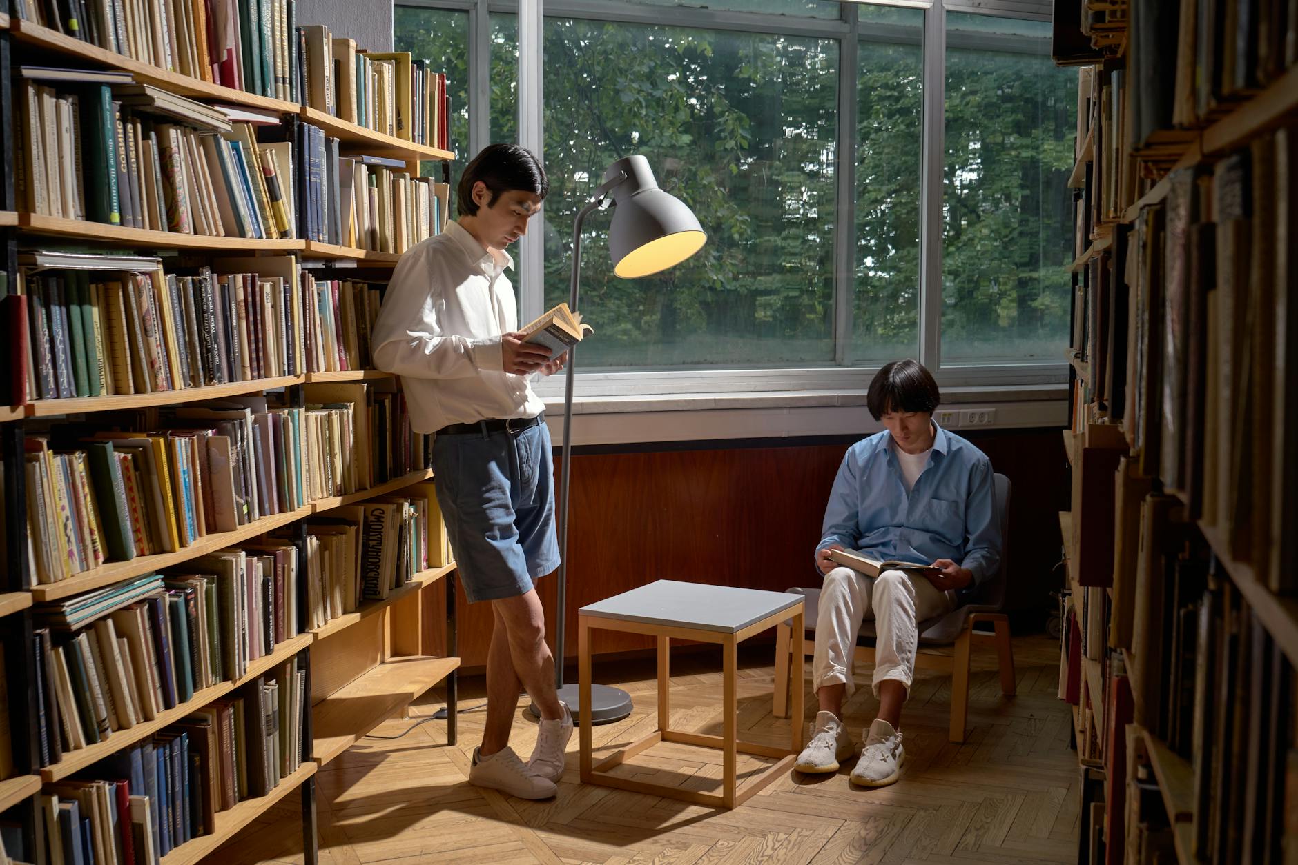 Two men reading in a cozy library setting, surrounded by bookshelves and natural light.