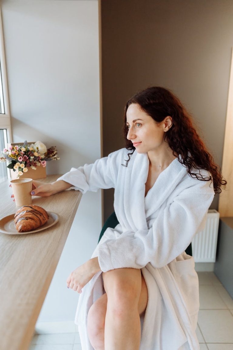 Young woman in a bathrobe enjoys a peaceful morning with coffee and croissant indoors.