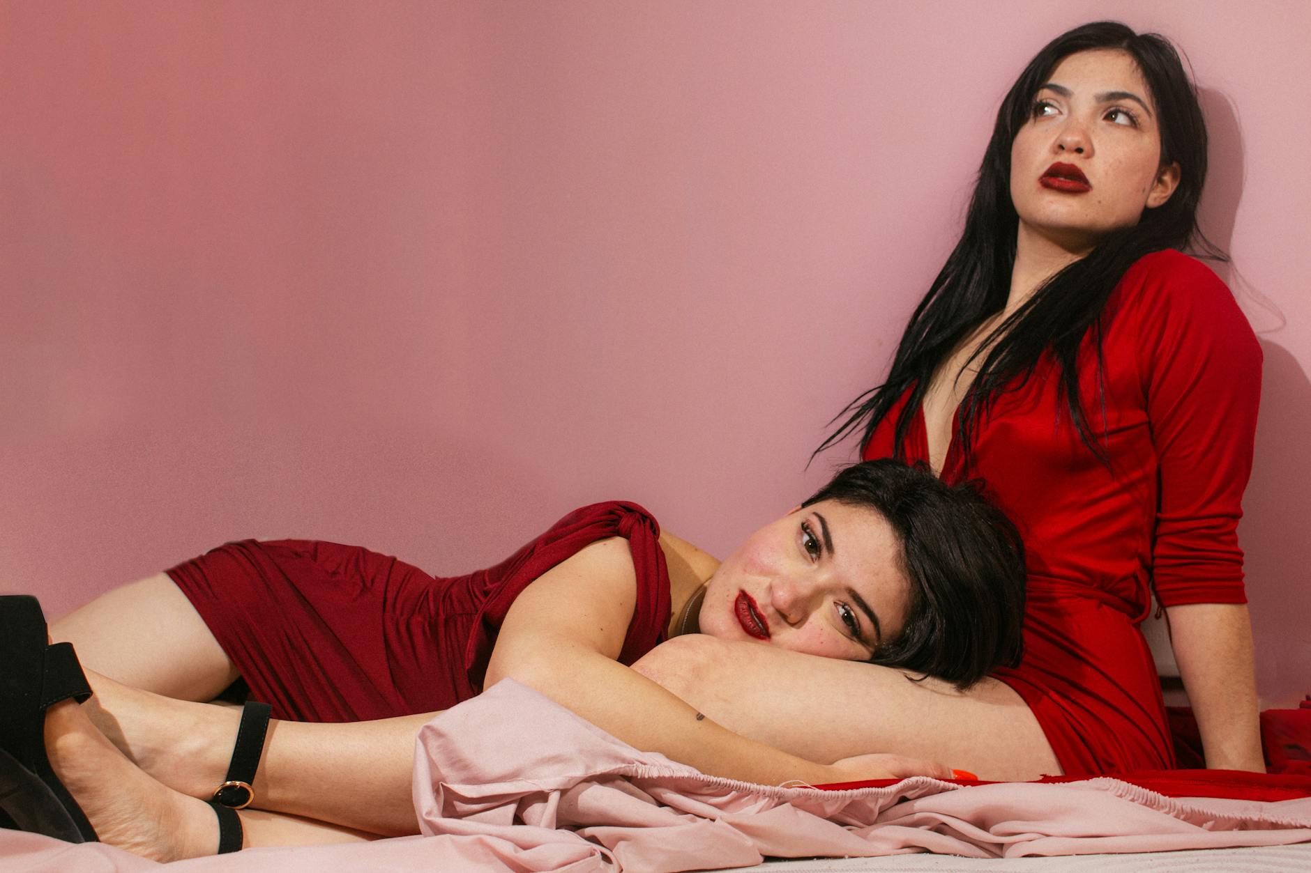 Two women posing in red dresses against a pink studio backdrop, capturing fashion and intimacy.