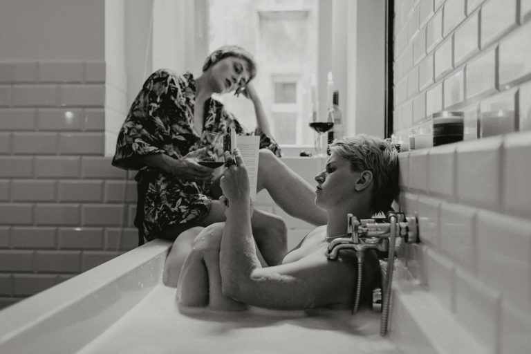 Two women enjoy a relaxing bath together, reading and sipping wine in a cozy atmosphere.