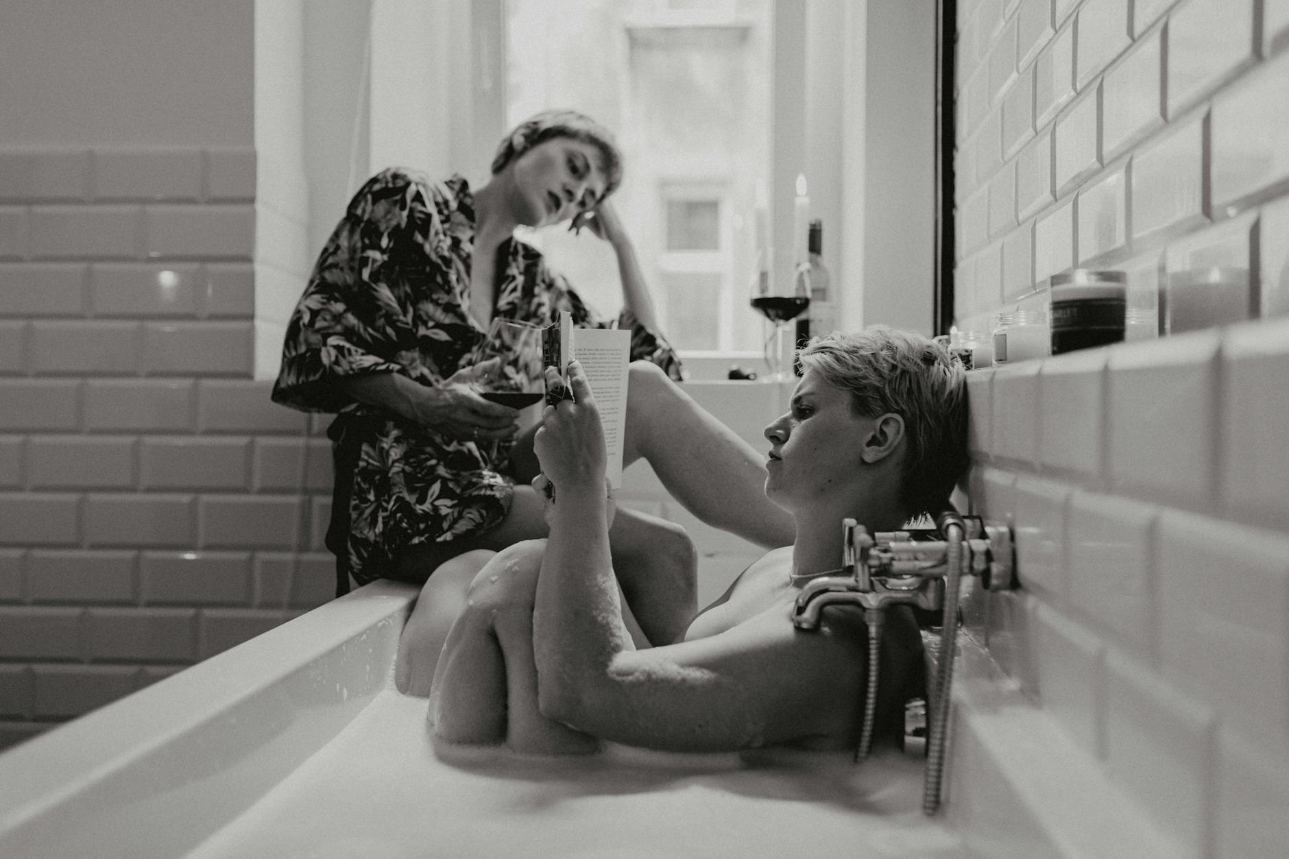 Two women enjoy a relaxing bath together, reading and sipping wine in a cozy atmosphere.