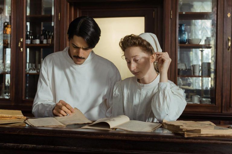 Two pharmacists in vintage attire examining old pharmaceutical texts in a historical pharmacy.