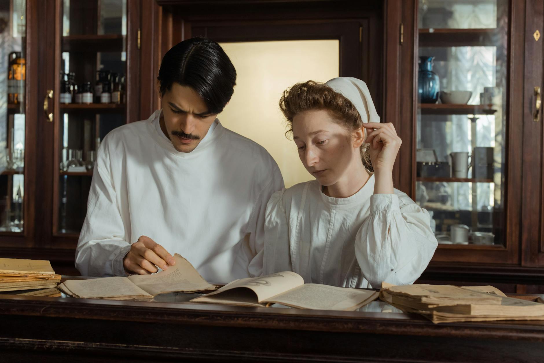 Two pharmacists in vintage attire examining old pharmaceutical texts in a historical pharmacy.