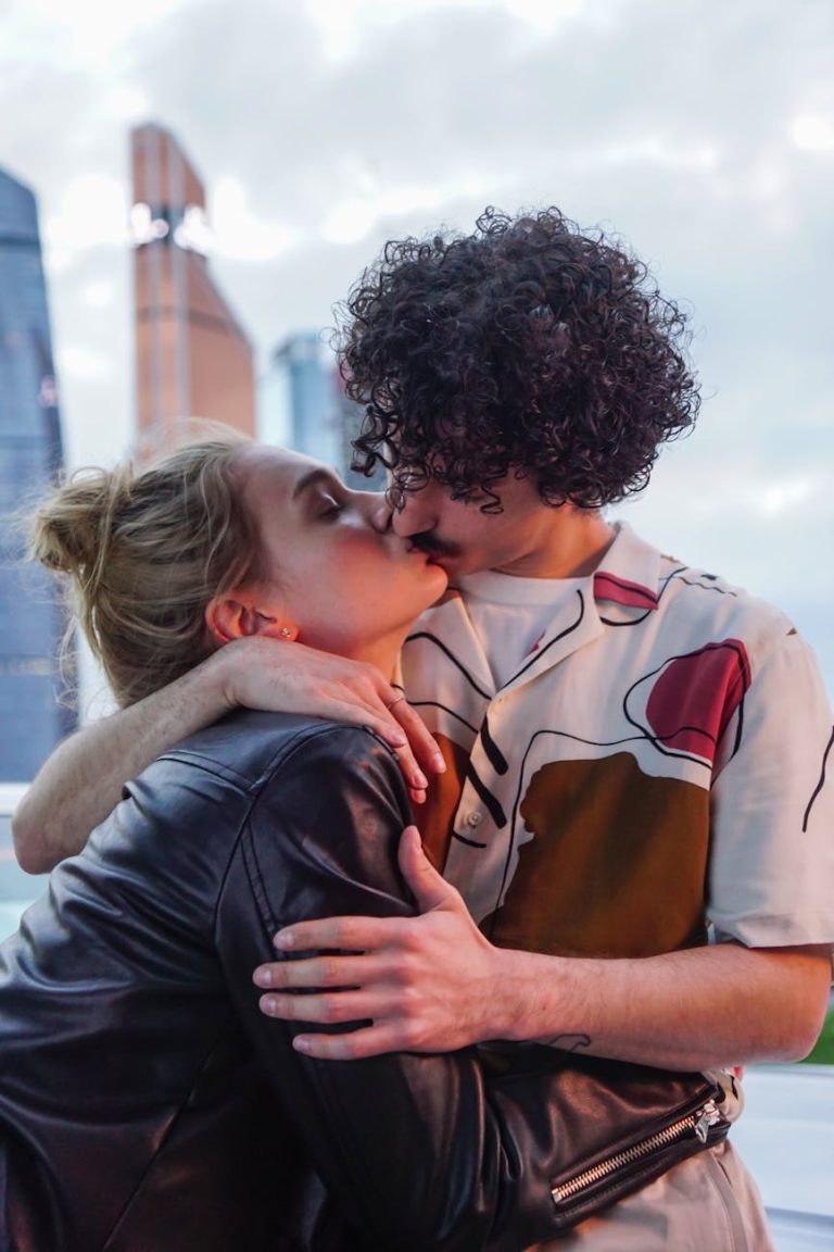 A romantic couple sharing a kiss outdoors at dusk with city skyscrapers in the background.