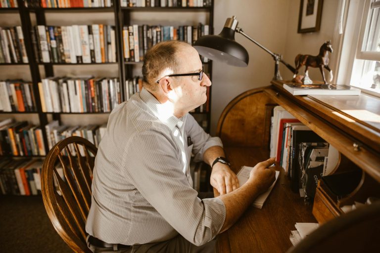 A man wearing glasses reads at his desk in a cozy home library filled with books.