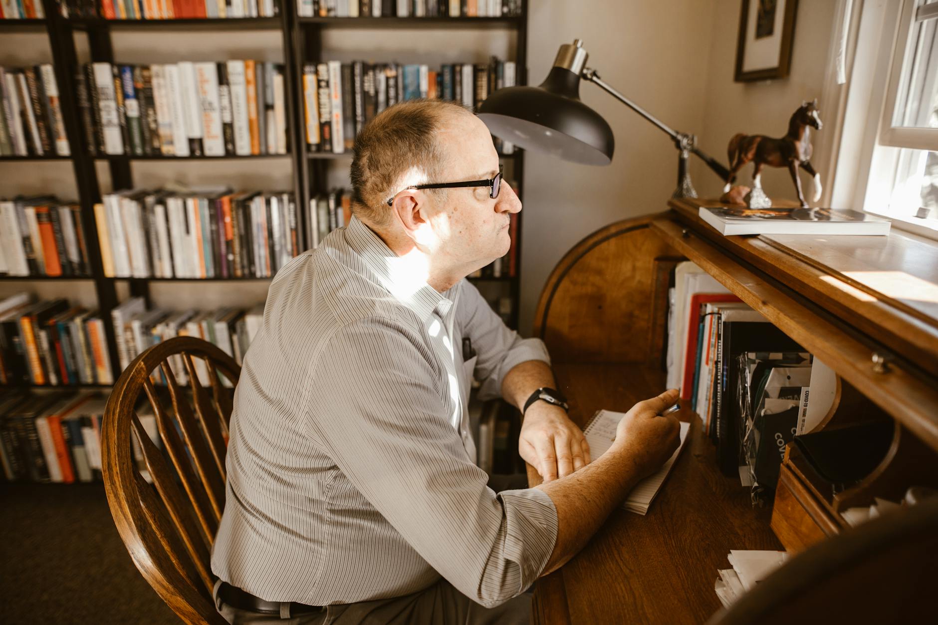 A man wearing glasses reads at his desk in a cozy home library filled with books.