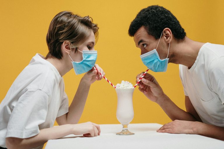 A couple wearing masks humorously shares a milkshake using straws over a white table.