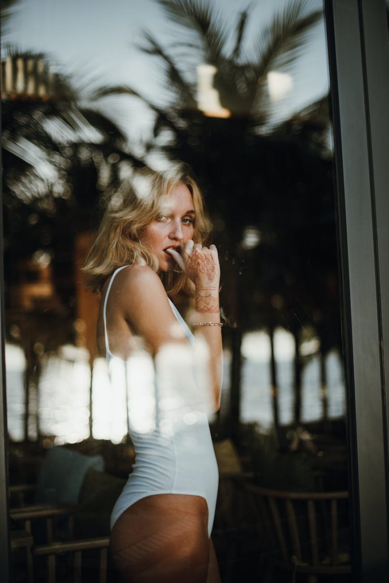 Blonde woman in a white swimsuit posing behind a glass pane, reflecting tropical surroundings.