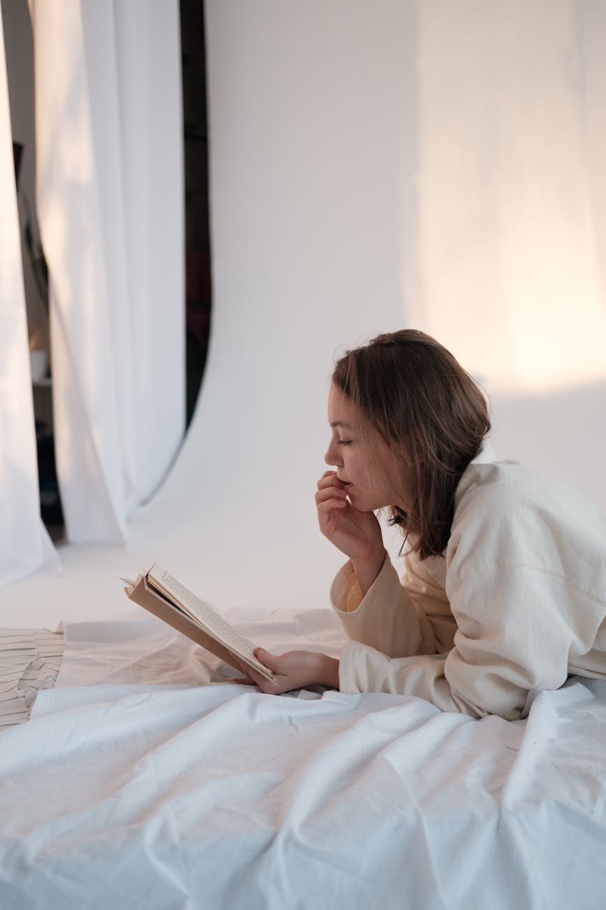 Side view of young woman touching face and reading interesting story while relaxing on mattress in morning