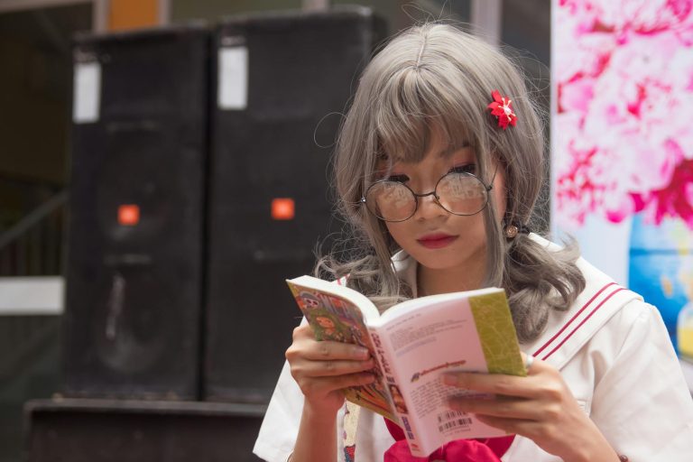 A young Asian woman in cosplay costume reads a book outdoors, wearing glasses.