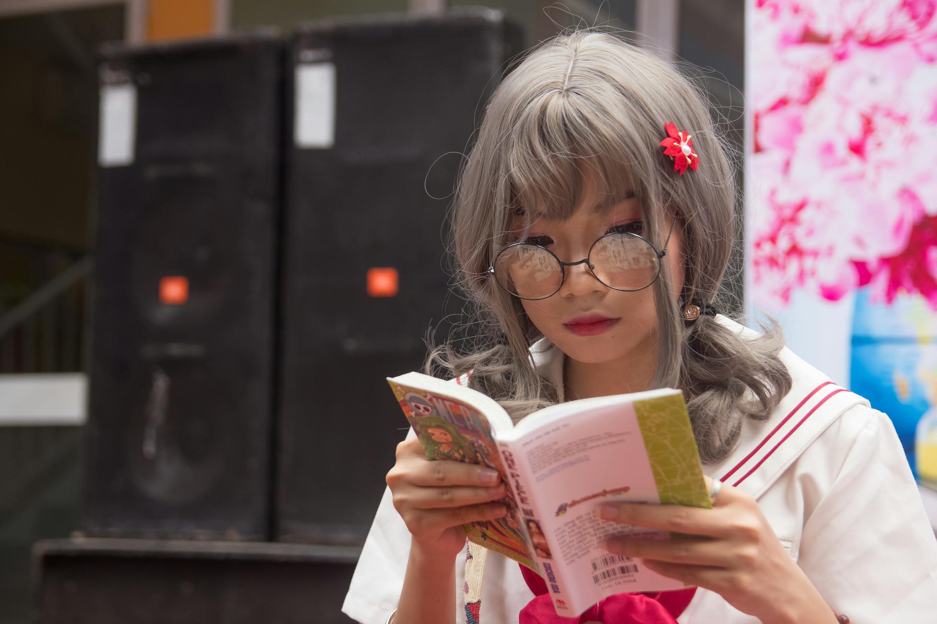 A young Asian woman in cosplay costume reads a book outdoors, wearing glasses.