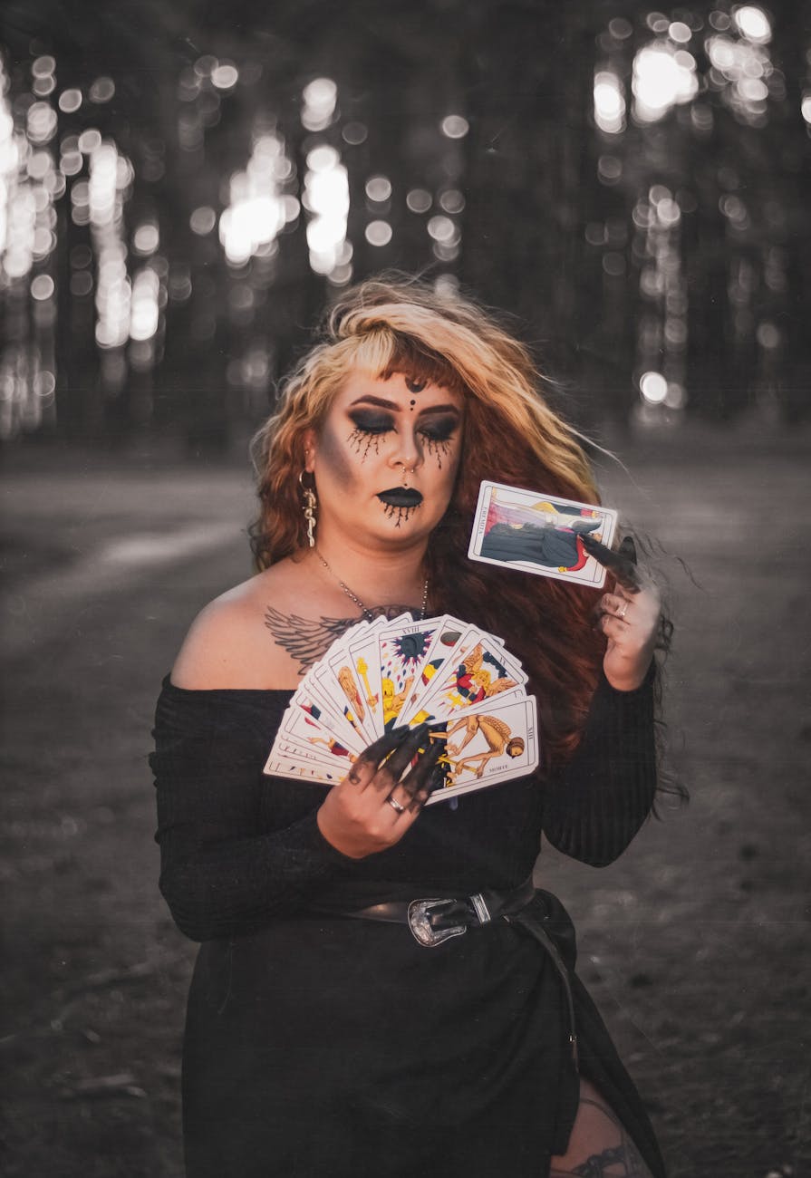 A mysterious woman with dark makeup and tattoos holds tarot cards in a forest, evoking a magical and eerie atmosphere.