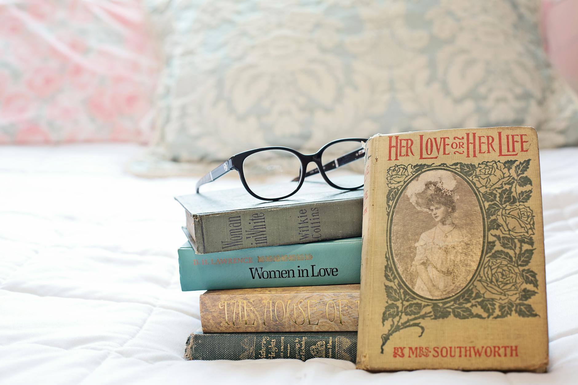 A stack of vintage romance novels with eyeglasses on a white bed.