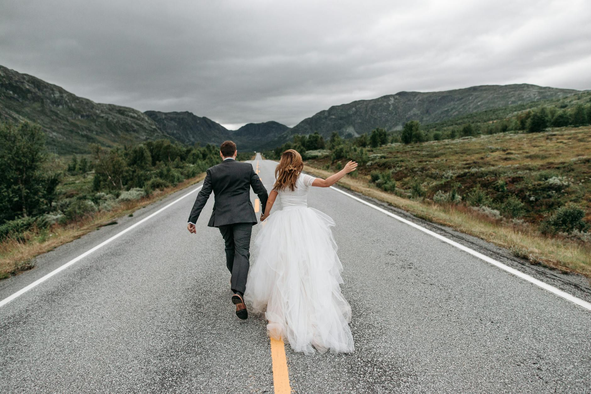 Bride and groom holding hands, running along a scenic mountain road. Capturing love and adventure.