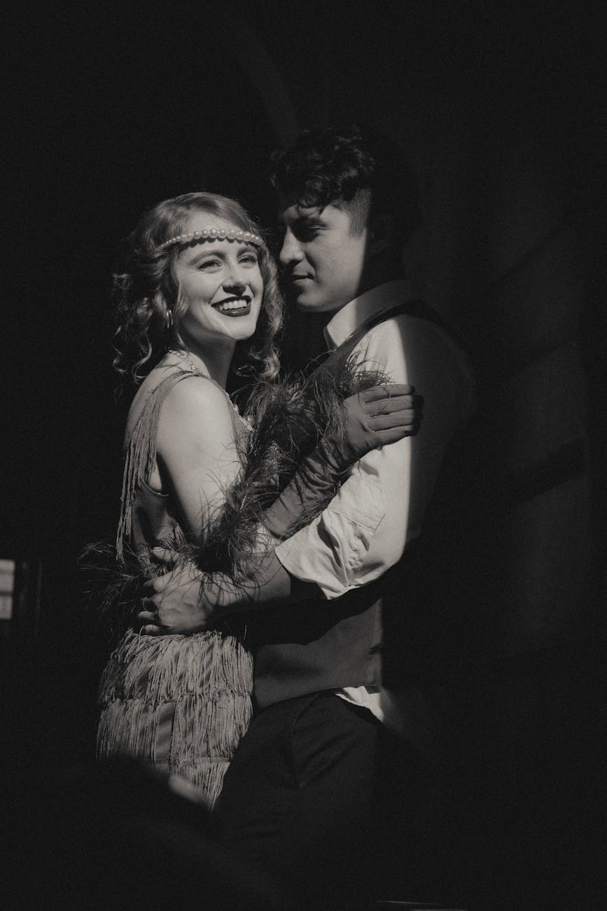 A smiling couple in vintage attire embracing in a dramatic black and white portrait.
