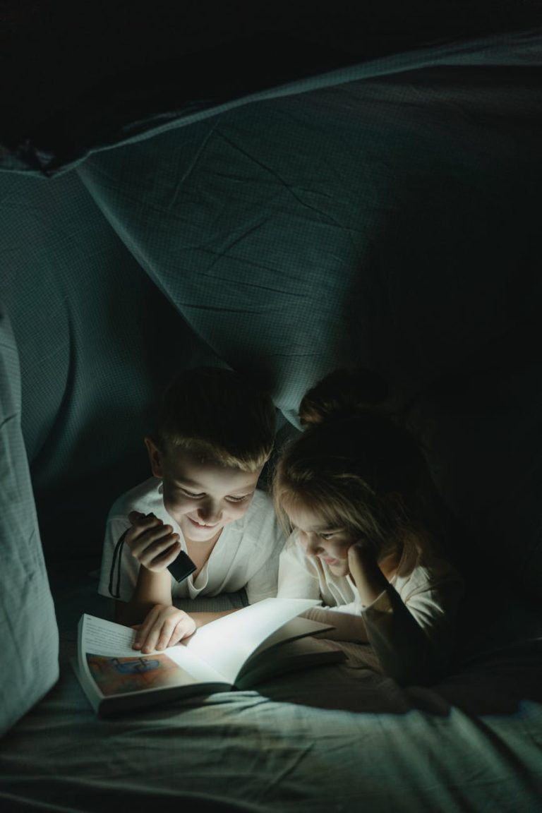Two children enjoy reading a book under a cozy blanket fort, creating a magical nighttime moment.