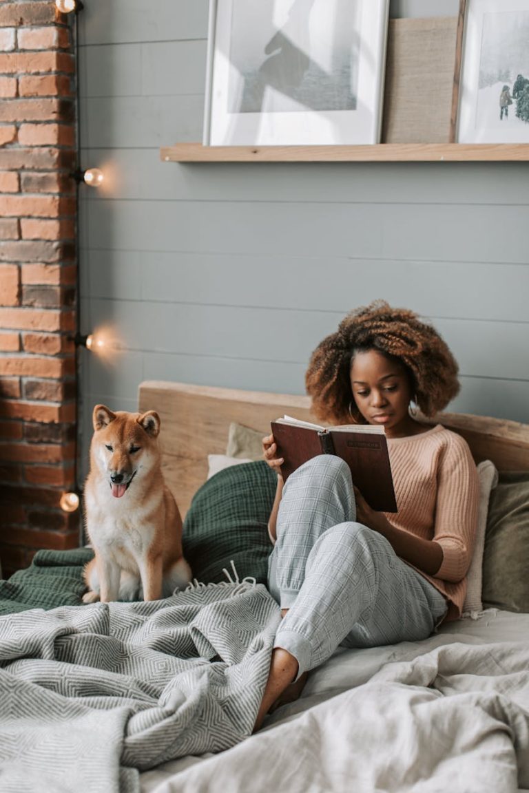 Young woman with afro hair reads a book in a cozy bedroom with her Shiba Inu dog.