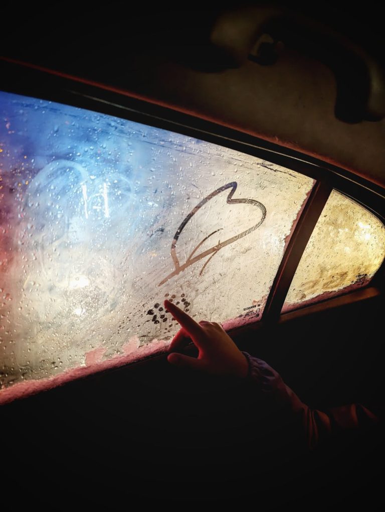 A hand draws a heart on a steamy car window with colorful light reflections.