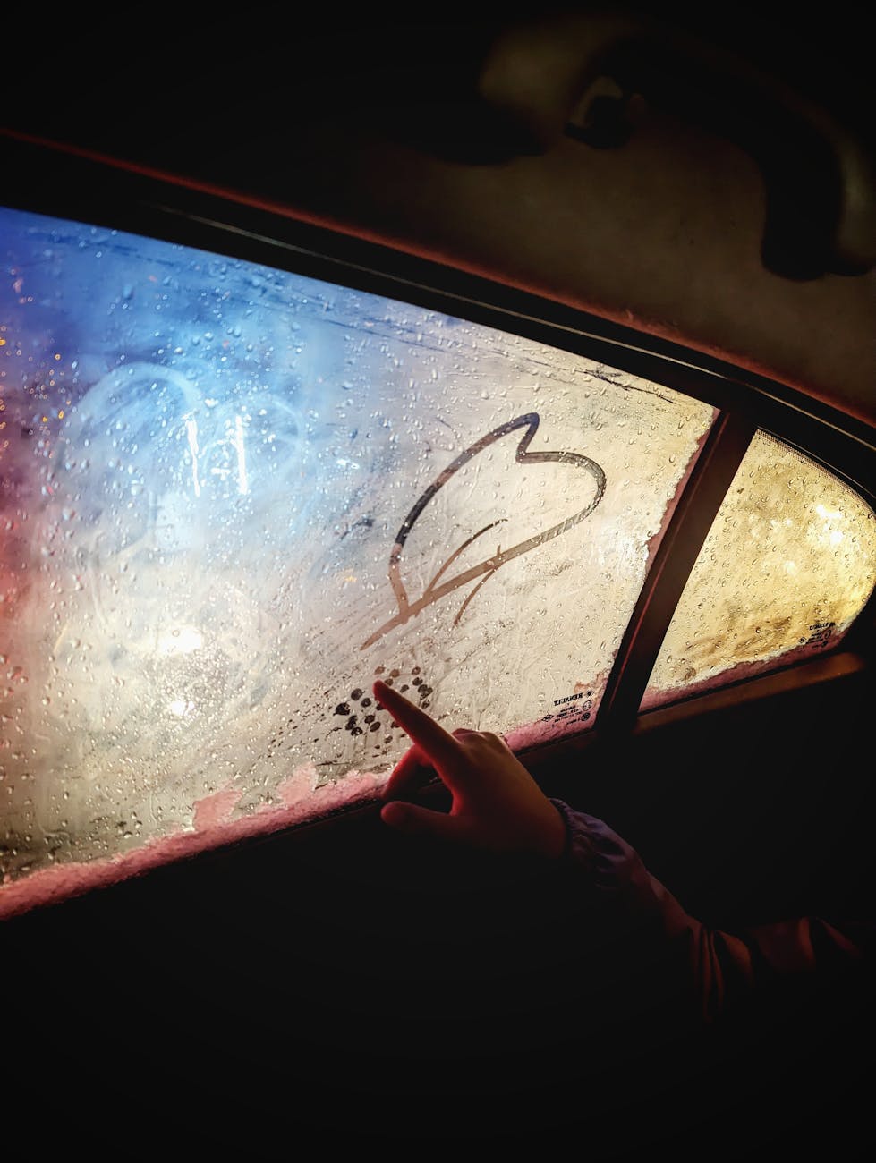 A hand draws a heart on a steamy car window with colorful light reflections.