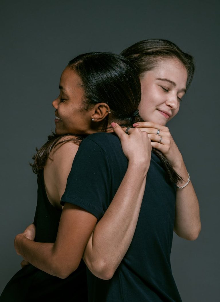 Two women of different ethnicities share a warm hug, symbolizing love and friendship against a gray backdrop.
